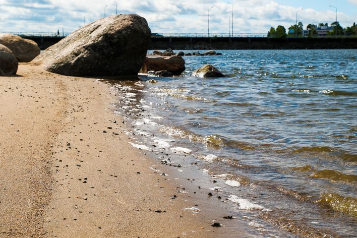 Strand med klippor vid vattnet och en bro i bakgrunden under en klar himmel.