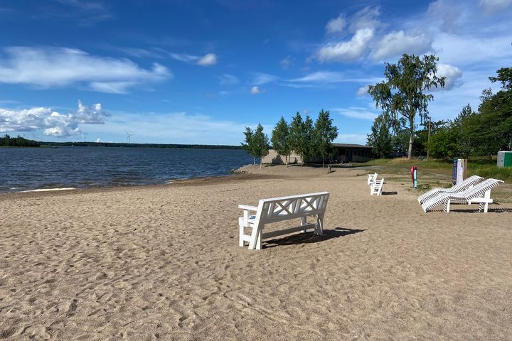 En solig sandstrand vid Gustavsborgs badstrand med vita bänkar och stolar.