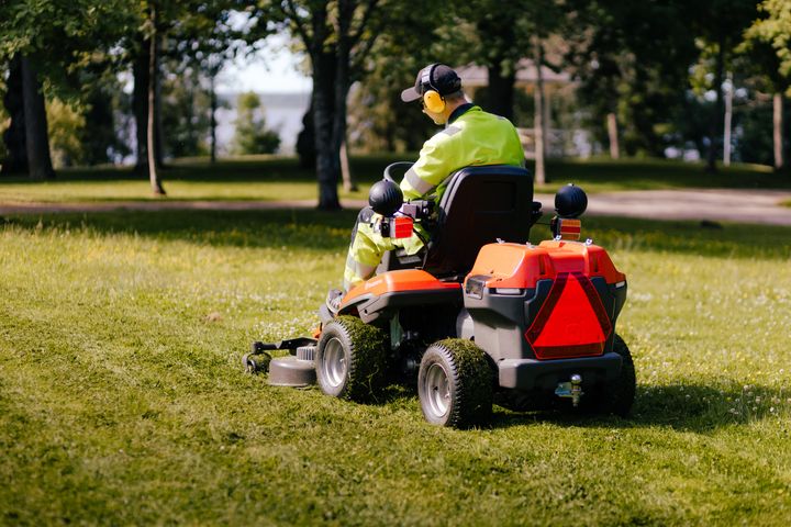 Henkilö ajaa päältä ajettavalla ruohonleikkurilla nurmikolla puistossa.