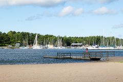 En strand med en brygga. I bakgrunden syns flera segelbåtar vid en småbåtshamn.