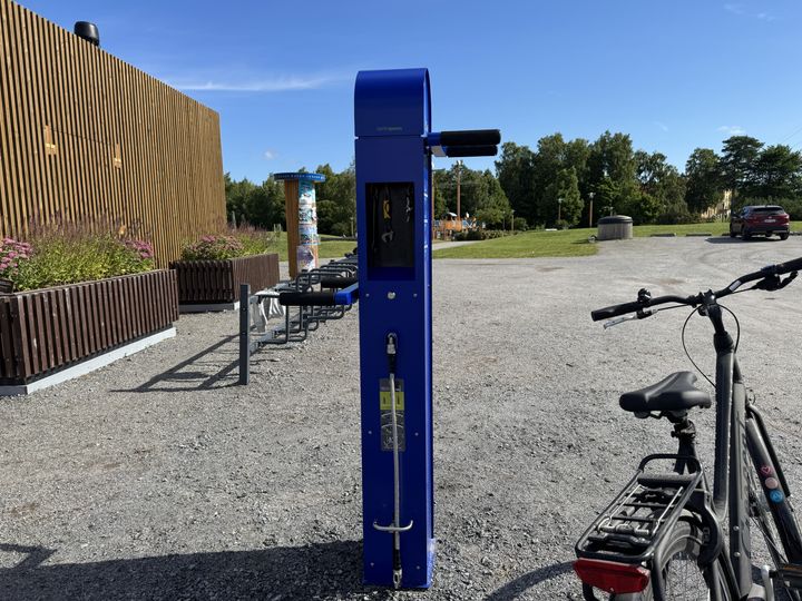 Sininen pyöränhuoltoteline Onkilahden kahvilan vieressä. Telineessä on syvennys, jonka sisällä on työkaluja, sekä molemmilla puolilla tangot, joihin voi ripustaa pyörän. Syvennyksen alapuolella on pyörän pumppu, joka toimii polkimella.