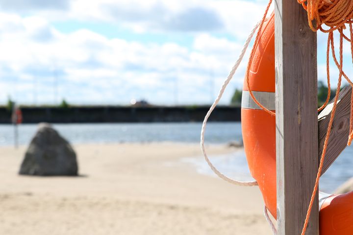 Orange livboj hänger på en stolpe vid en sandstrand med vatten i bakgrunden.