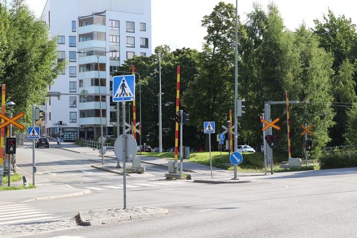 Konepajakadun tasoristeys, jossa on liikennevalot ja tasoristeysmerkki. Taustalla valkoinen kerrostalo ja puita.