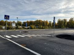 Tyhjä pysäköintialue, suojatie ja liikennemerkkejä syksyisten puiden edessä.