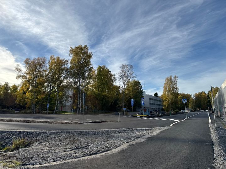 Syksyinen katunäkymä: puiden reunustama tie, uusi asfaltti ja rakennuksia taustalla.