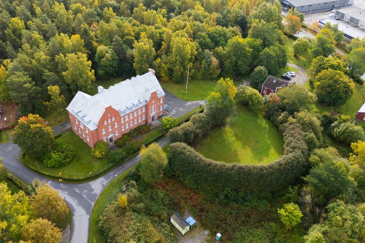 Ilmakuva punaisesta tiilirakennuksesta metsän keskellä, vieressä pensasaidan ympäröimä nurmikenttä.