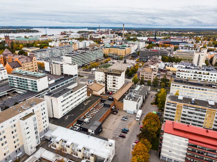 Flygbild över stadskärna med byggnader, en parkeringsplats och vattnet synligt i bakgrunden.