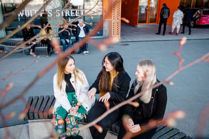 Kolme naispuolista VAMKin opiskelijaa istuu ja keskustelee ulkona, taustalla näkyy Wollfintien kampuksen kahvilan kyltti ja muita opiskelijoita.