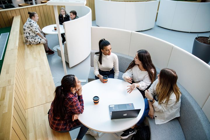 Neljä henkilöä istuu keskustelemassa pöydän ääressä modernissa korkeakouluympäristössä. Taustalla muita opiskelijoita.