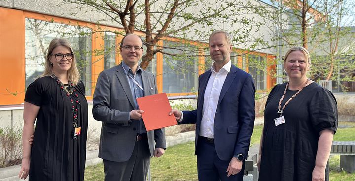Neljä hymyilevää henkilöä seisoo Vaasan ammattikorkeakoulun sisäpihalla, kaksi naista ja kaksi miestä, joista yksi pitää kädessään oranssia kansioita. Taustalla näkyy VAMKin päärakennus ja vihreitä puita.