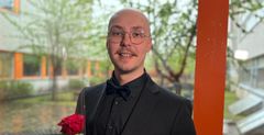 Arsi Ollila, VAMKin insinööriopiskelija, seisoo ikkunan edessä taustalla VAMKin sisäpiha yllään musta puku ja rusetti, pitäen ruusua kädessään.