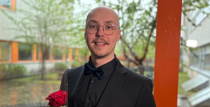 Arsi Ollila, VAMKin insinööriopiskelija, seisoo ikkunan edessä taustalla VAMKin sisäpiha yllään musta puku ja rusetti, pitäen ruusua kädessään.