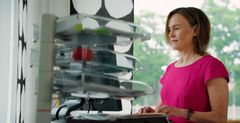 A woman in a pink shirt stands at a desk while working on a computer.