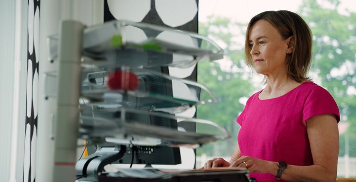 A woman in a pink shirt stands at a desk while working on a computer.