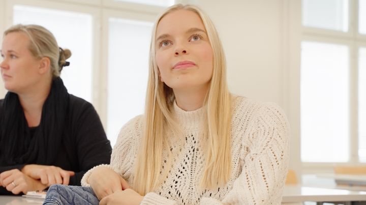 Kaksi henkilöä istuu luokkahuoneessa, katsellen eteenpäin.