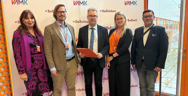 Viisi henkilöä seisoo yhdessä sisätiloissa VAMK-logolla varustetun taustan edessä.