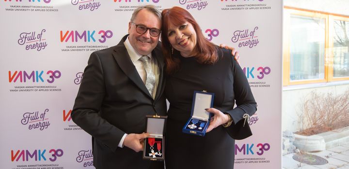 Kaksi henkilöä hymyilee ja poseeraa yhdessä kunniamerkkien kanssa VAMK-taustaseinän edessä.