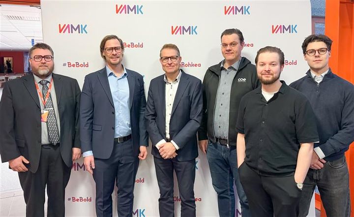 Alt-teksti: Kuusi miestä seisoo rivissä VAMKin logolla varustetun taustaseinän edessä.