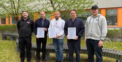 Five people standing outdoors, three holding certificates, with a building in the background.