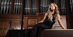 Alumna of the year sits confidently in front of a large pipe organ, wearing a black gown, in a wood-paneled hall