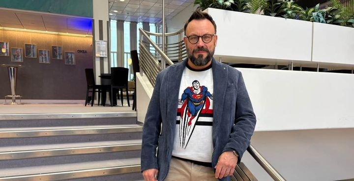 Riku Rantala seisoo portaikon vierellä sisätiloissa. Hänellä on sininen bleiseri Superman-aiheisen t-paidan päällä ja beigit housut.