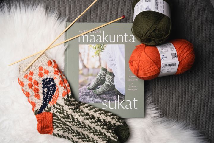 Taljalle aseteltuina Yhteishyvän maakuntasukat -kirja, valmiit neulotut Satakunta-sukat, lankaa ja neulepuikot