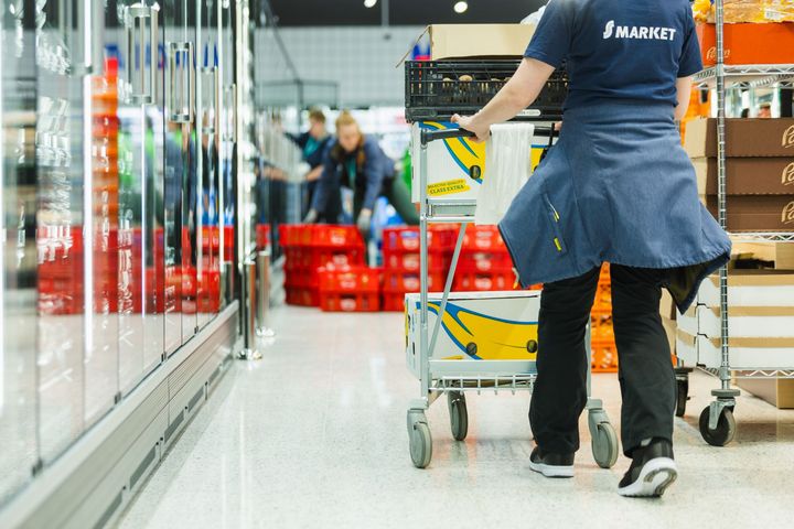 Alt-teksti: Työntekijä kaupan käytävällä työntämässä kärryä, jossa on tavaralaatikoita.