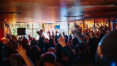 Joukko ihmisiä nauttimassa live-esityksestä ravintolassa, kädet ilmassa ja värikkäät valot taustalla.