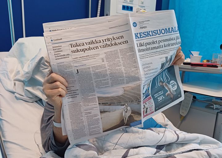 Henkilö lukee sanomalehteä sairaalasängyssä.