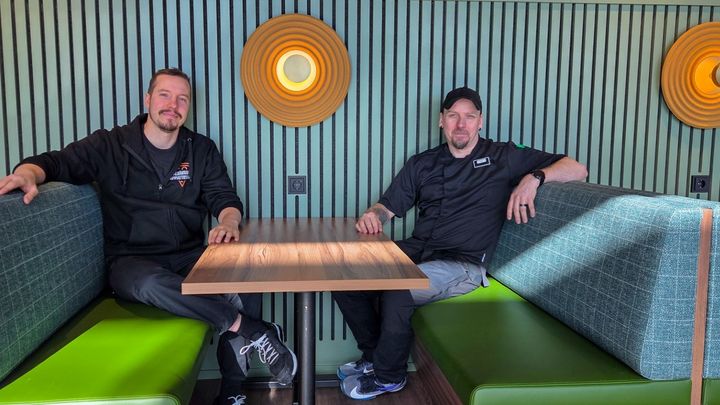 Kaksi miestä istuu ravintolan vihreillä penkeillä pöydän ääressä. Taka-alalla on pyöreitä valaisimia.