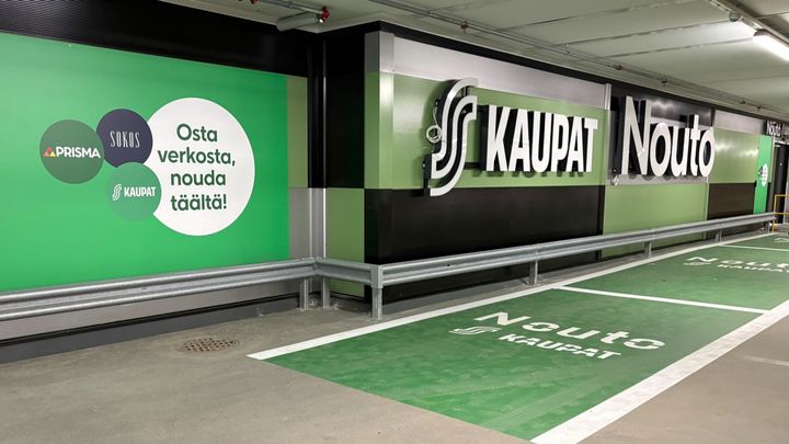 Alt-teksti: Prisman noutopiste parkkihallissa, jossa on vihreitä opasteita ja mainoksia verkkokaupan käytöstä.