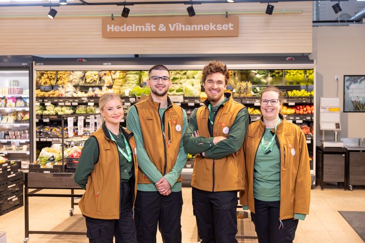 Neljä työntekijää seisoo hymyillen hedelmä- ja vihannesosaston edessä.