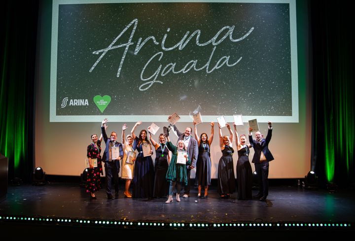 Ryhmä ihmisiä poseeraa lavalla "Arina Gaala" -tapahtumassa, pidellen diplomeja iloisina.