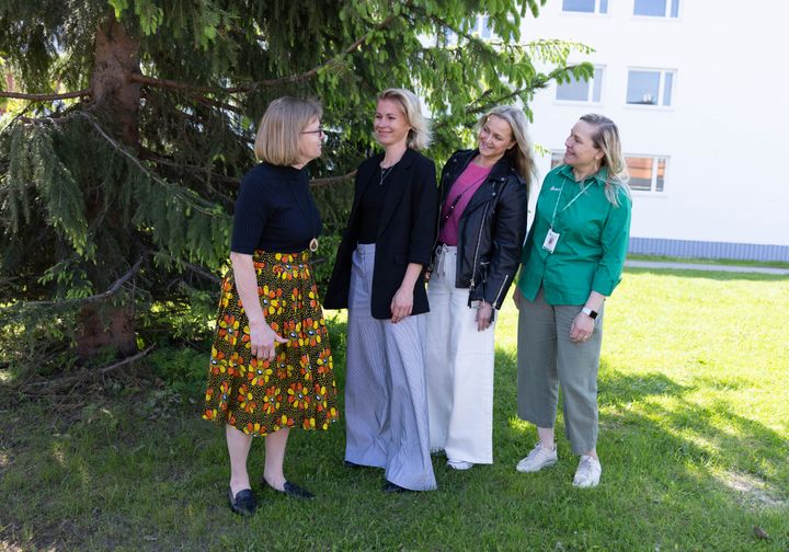 Kuvassa on neljä henkilöä seisomassa ulkona ruohikolla. Taustalla on kuusi ja valkoinen rakennus.