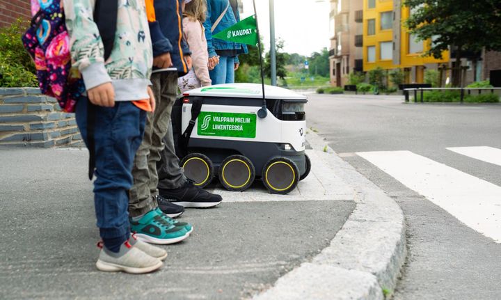 Lapset seisovat suojatien reunalla yhdessä pienen toimitusrobotin kanssa.
