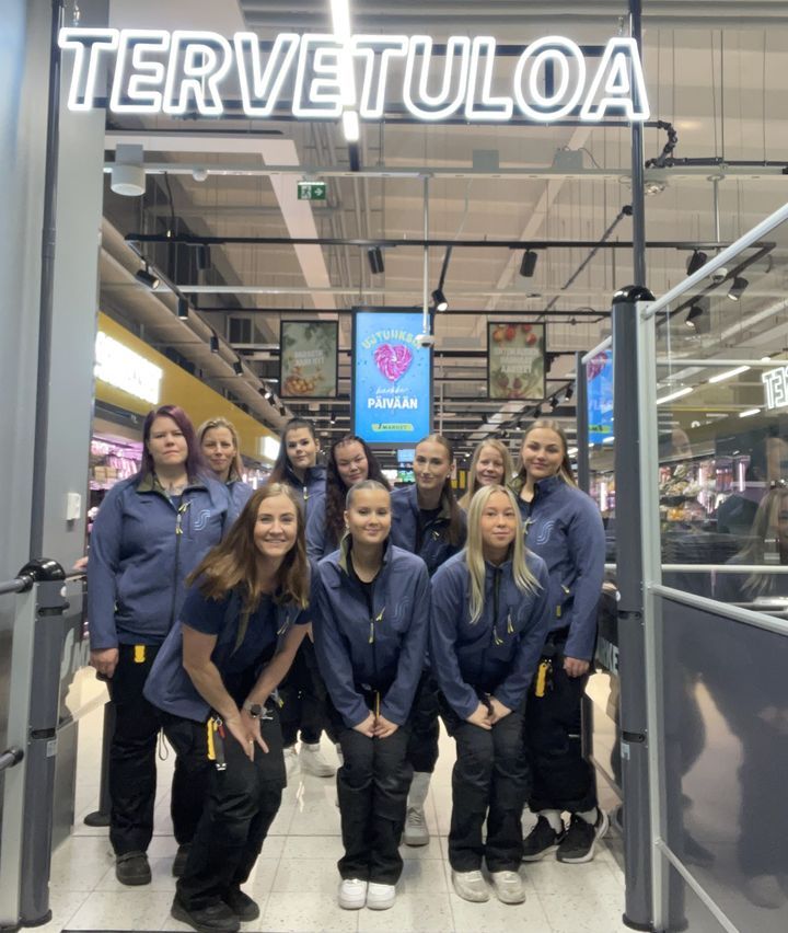 Ryhmä ihmisiä seisoo kaupassa "Tervetuloa"-kyltin alla.