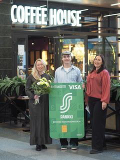 Kolme henkilöä seisoo Coffee House -kahvilan edessä. Keskimmäinen henkilö pitelee suurta vihreää korttia, ja vasemmanpuoleinen henkilö pitelee kukkakimppua.