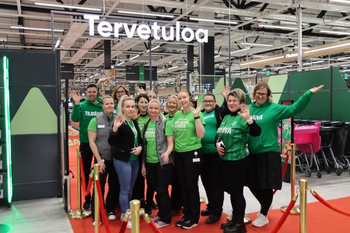 Ryhmä iloisia ihmisiä tervehtii kameroita supermarketissa, jossa taustalla lukee "Tervetuloa".