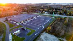 Ilmakuva suuresta, parkkipaikkojen ympäröimästä liiketilasta maaseudun maisemassa. Aurinko paistaa matalalta taustalla.