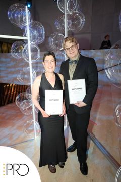 Kaksi henkilöä seisoo rinnakkain ja pitelee diplomeja juhlallisessa tilaisuudessa.