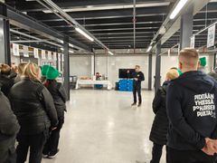 Ryhmä ihmisiä seisoo sisätiloissa kuuntelemassa puhujaa. Taustalla on pöytiä, joilla on tarjottavia ja juomia.