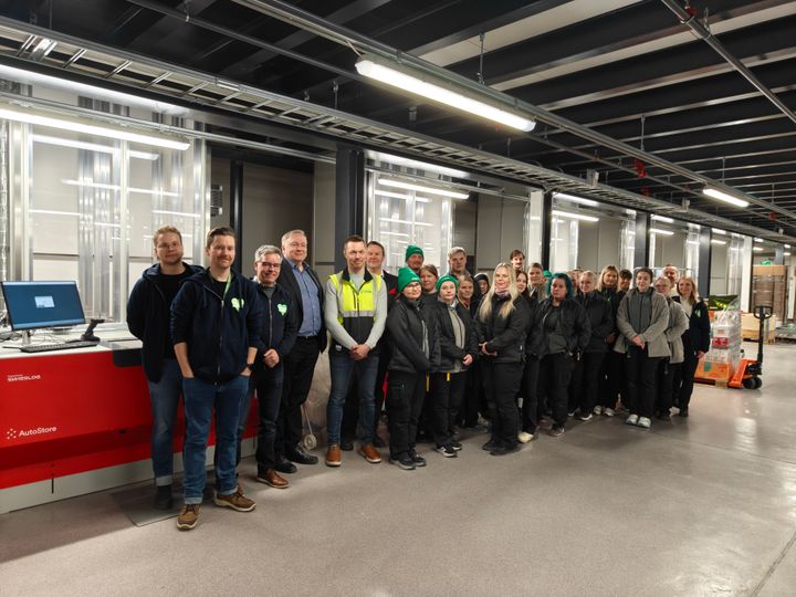 Ryhmä ihmisiä seisoo rivissä sisätiloissa. Taustalla näkyy valaistuja laatikoita ja tietokone.