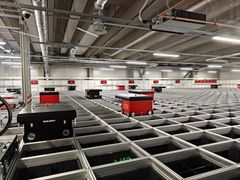 Automaattinen varastojärjestelmä, jossa punaisia kuljetusrobotteja liikkuu metallikourujen yläpuolella.