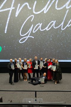 Ryhmä ihmisiä seisoo lavalla ja pitelee palkintoja. He ovat pukeutuneet juhlavasti. Taustalla lukee "Arinan Gaala".