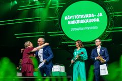 Henkilöt seisovat vihreällä lavalla ja vastaanottavat palkintoja. Taustalla on suuri, pyöreä näyttö, jossa lukee "Käyttötavara- ja erikoiskauppa".