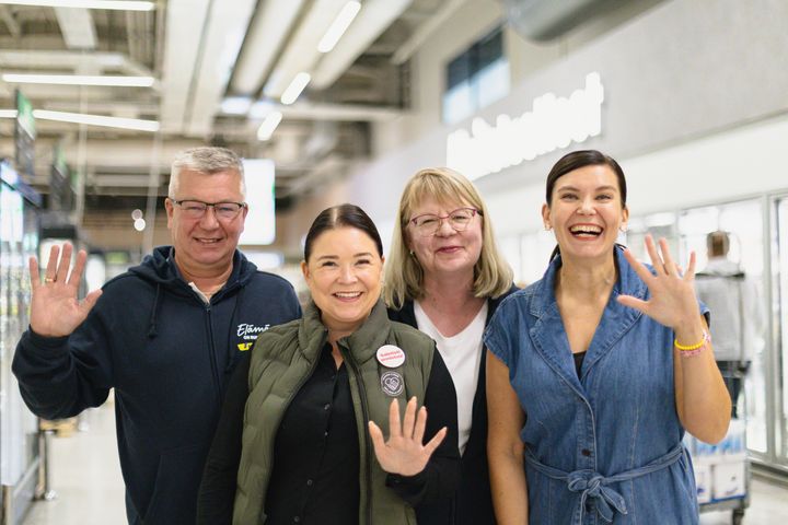 Neljän iloisesti hymyilevän henkilön ryhmä sisätiloissa, kaikki heiluttavat kättään kameralle.