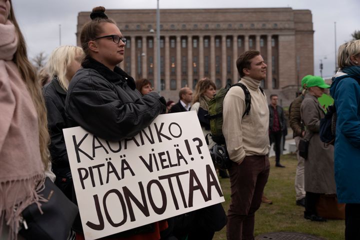 Mielenosoitus eduskuntatalon edessä, mielenosoittajalla kyltti Kauanko pitää vielä jonottaa.