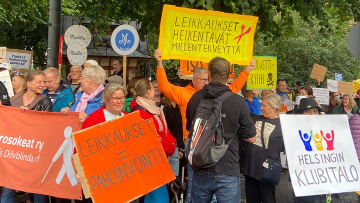 Mielenosoittajia kylttien kanssa, kylteissä leikkauksia vastustavia ja mielenterveyttä puolustavia sloganeita.