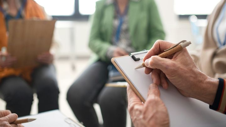 Lähikuva lehtiöön kirjoittavista käsistä kokoustilanteessa muiden osittain näkyvien läsnäolijoiden kanssa.