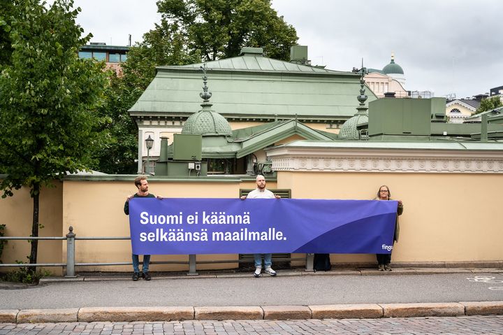 Kuvassa sinisävyinen banderolli, jossa teksti: Suomi ei käännä selkäänsä maailmalle.
